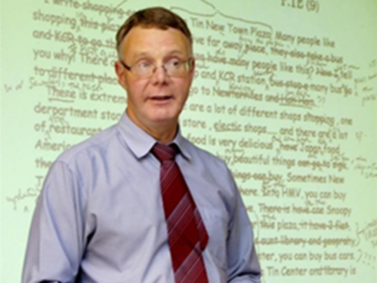 Neil MacKay, in a shirt and tie whilst standing and teaching in front of a whiteboard.