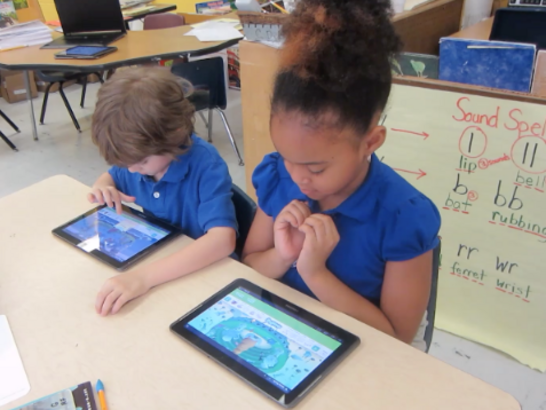 Two children sit together in their school uniforms, both playing Nessy on their tablets.
