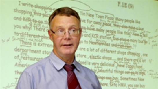 Neil MacKay, in a shirt and tie whilst standing and teaching in front of a whiteboard.